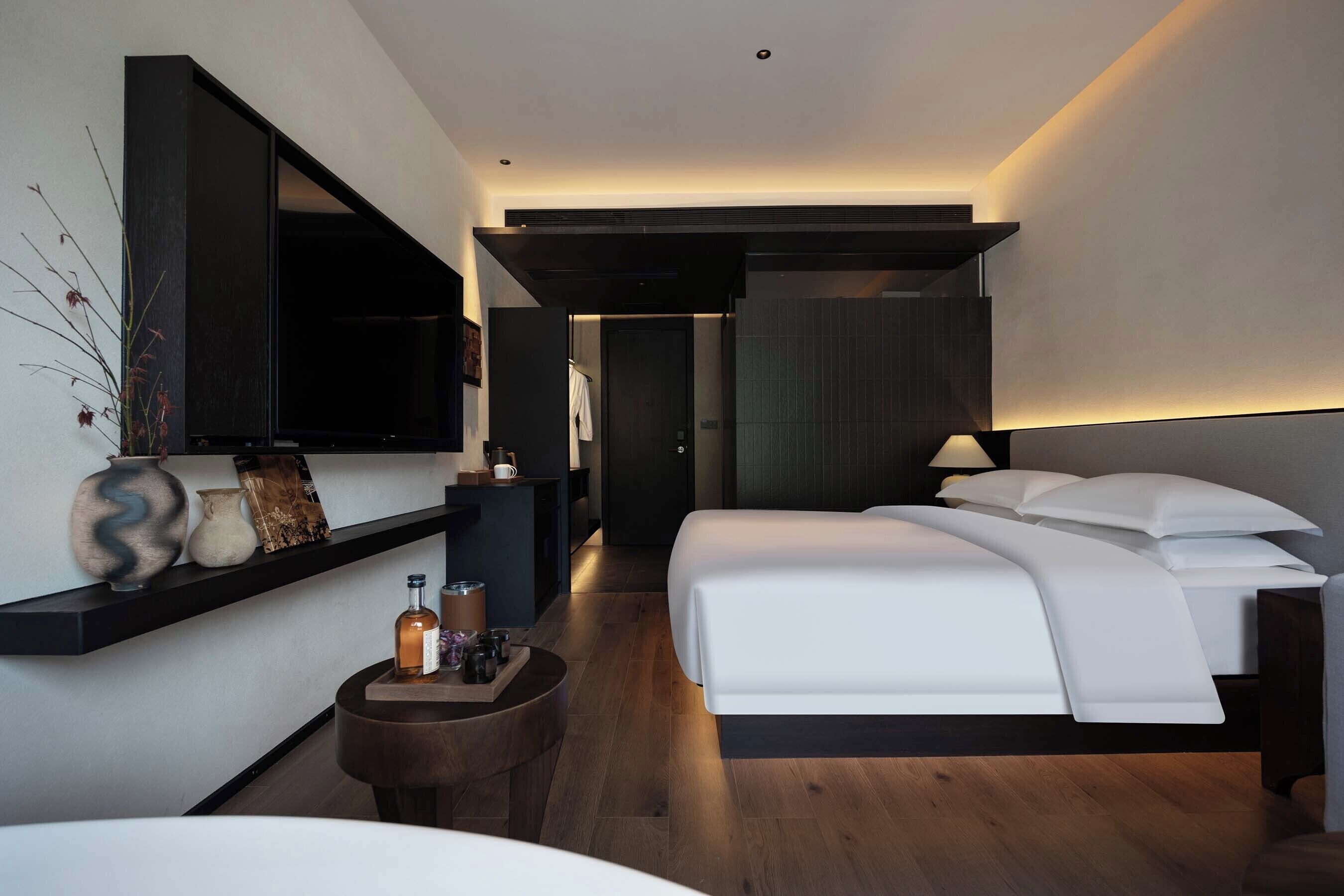 A modern hotel room with a large bed, a bedside table, a wall-mounted TV, and a dark wooden floor.