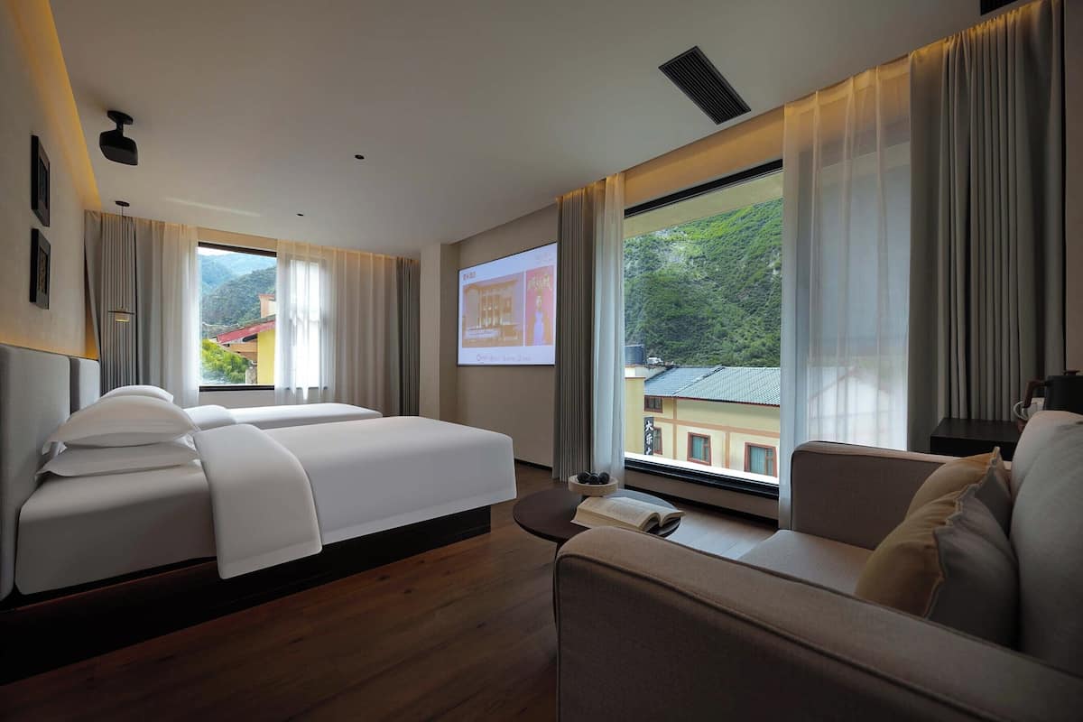 A hotel room with two beds, a sofa, a projector screen, and a view of a building and greenery.