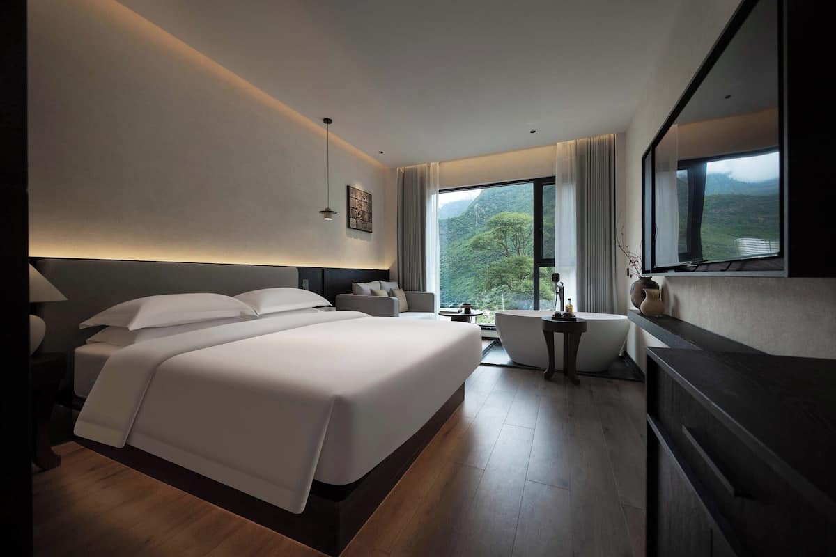 A modern hotel room with a large bed, a desk, a sofa, and a view of the outdoors.