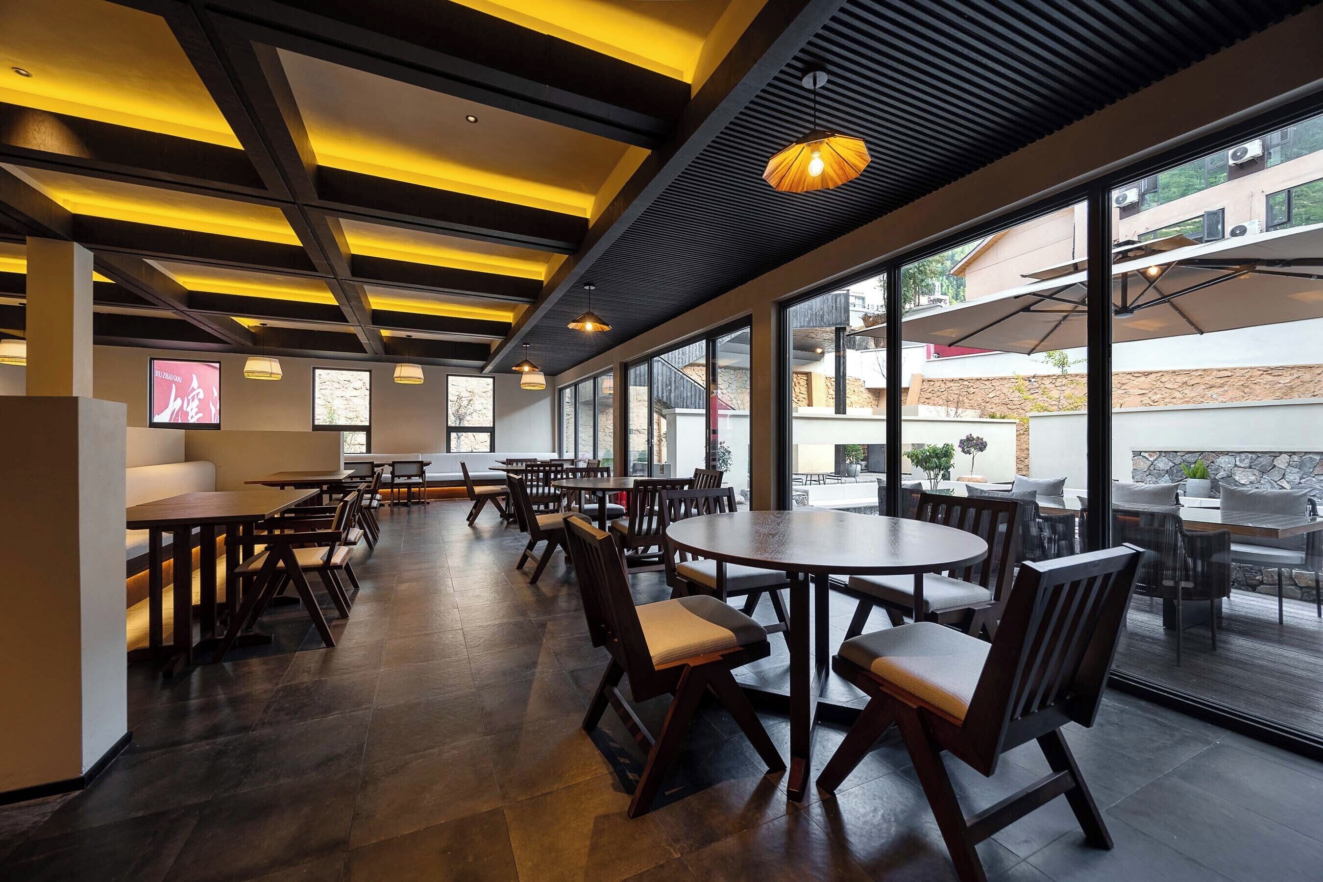 A modern restaurant with wooden tables and chairs, large windows, and a ceiling with exposed beams and lighting fixtures.