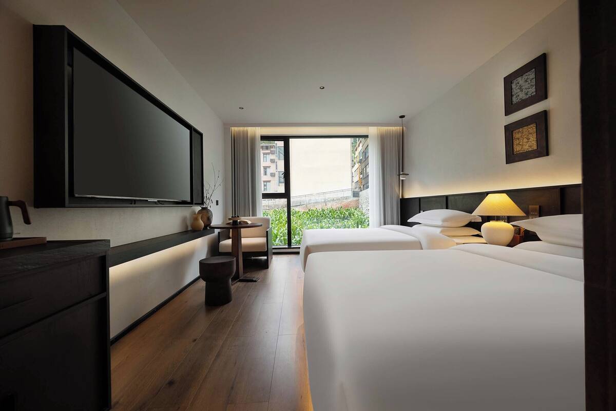 A hotel room with a large TV, two beds, a desk, a chair, a lamp, and a view of greenery outside.