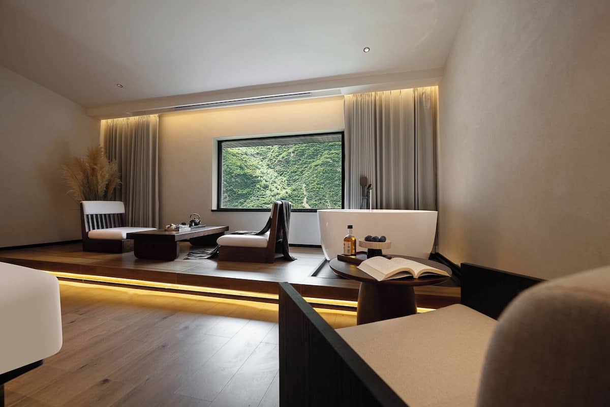 A modern room with a large window, a sofa, a coffee table, and a side table with a book and a bottle.