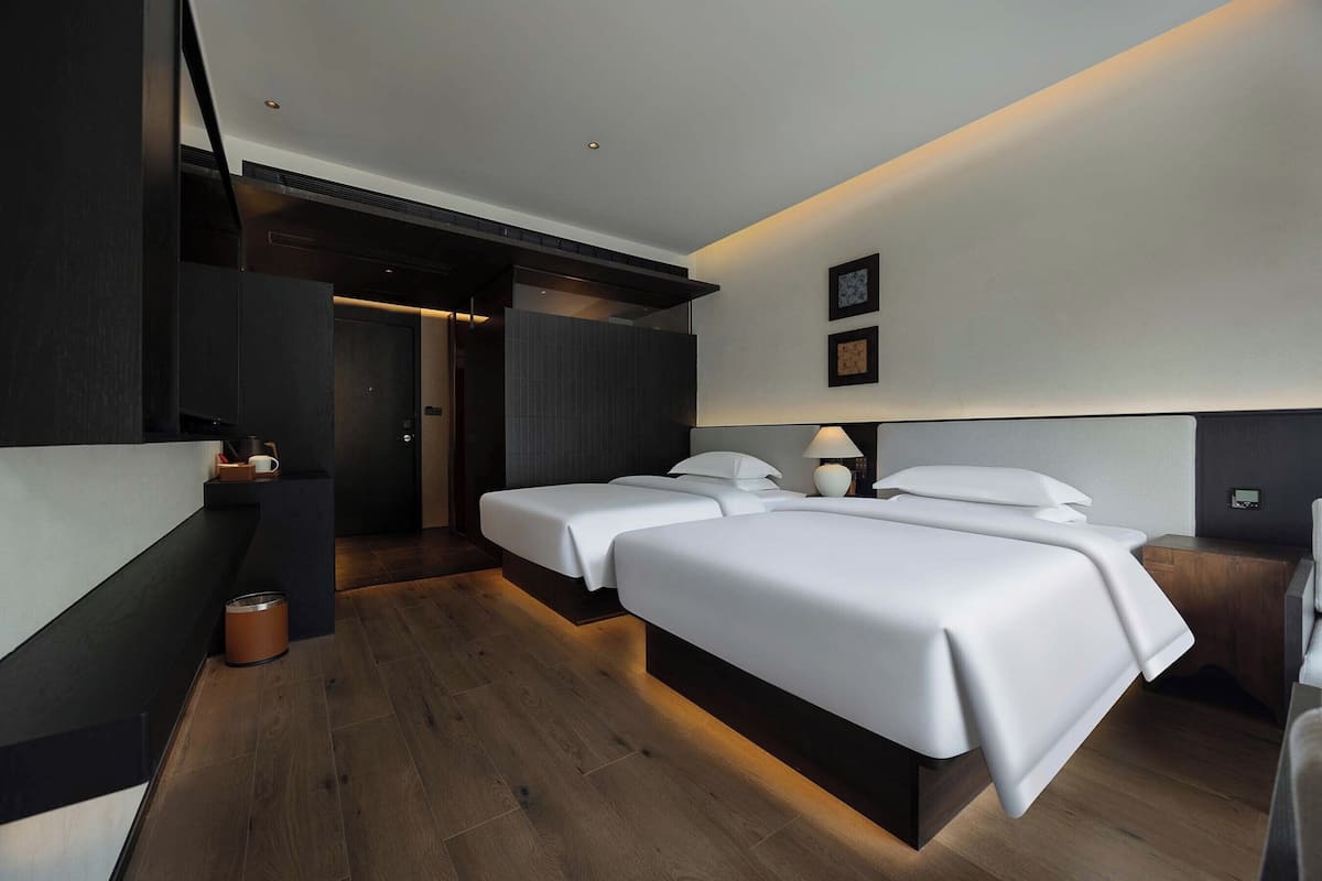A modern hotel room with two beds, a desk, a chair, a small table, and a trash bin.