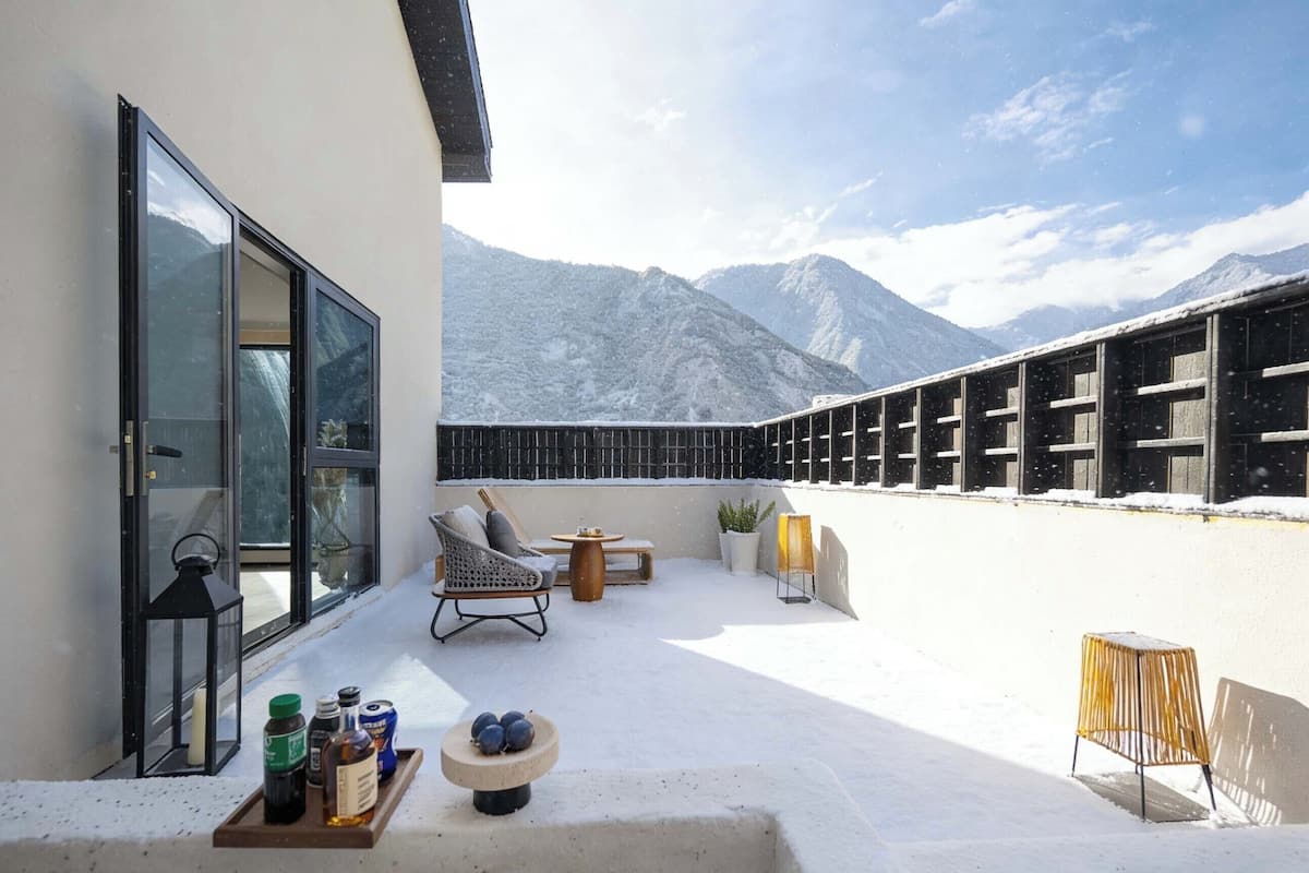 A balcony with a view of snow-covered mountains, a seating area with a table and chairs, and a small table with bottles and a bowl of fruit.