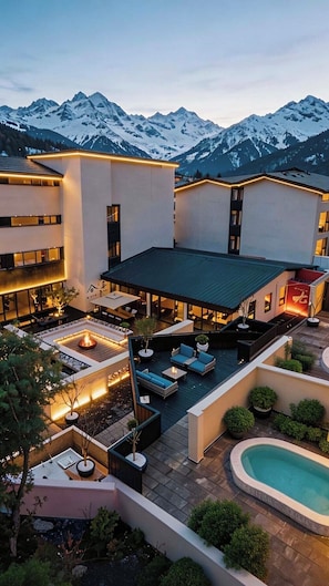 Une cour d’hôtel avec des espaces de repos en plein air, une piscine et, en arrière-plan, des montagnes enneigées.
