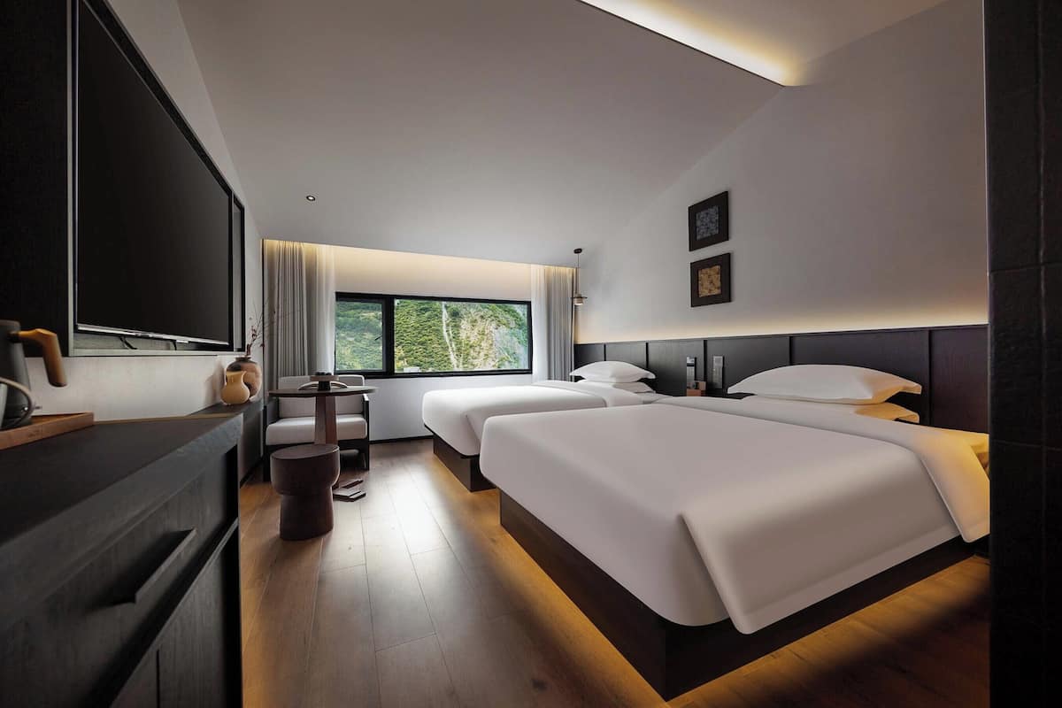 A hotel room with two beds, a desk, a chair, a TV, and a window with a view of greenery.