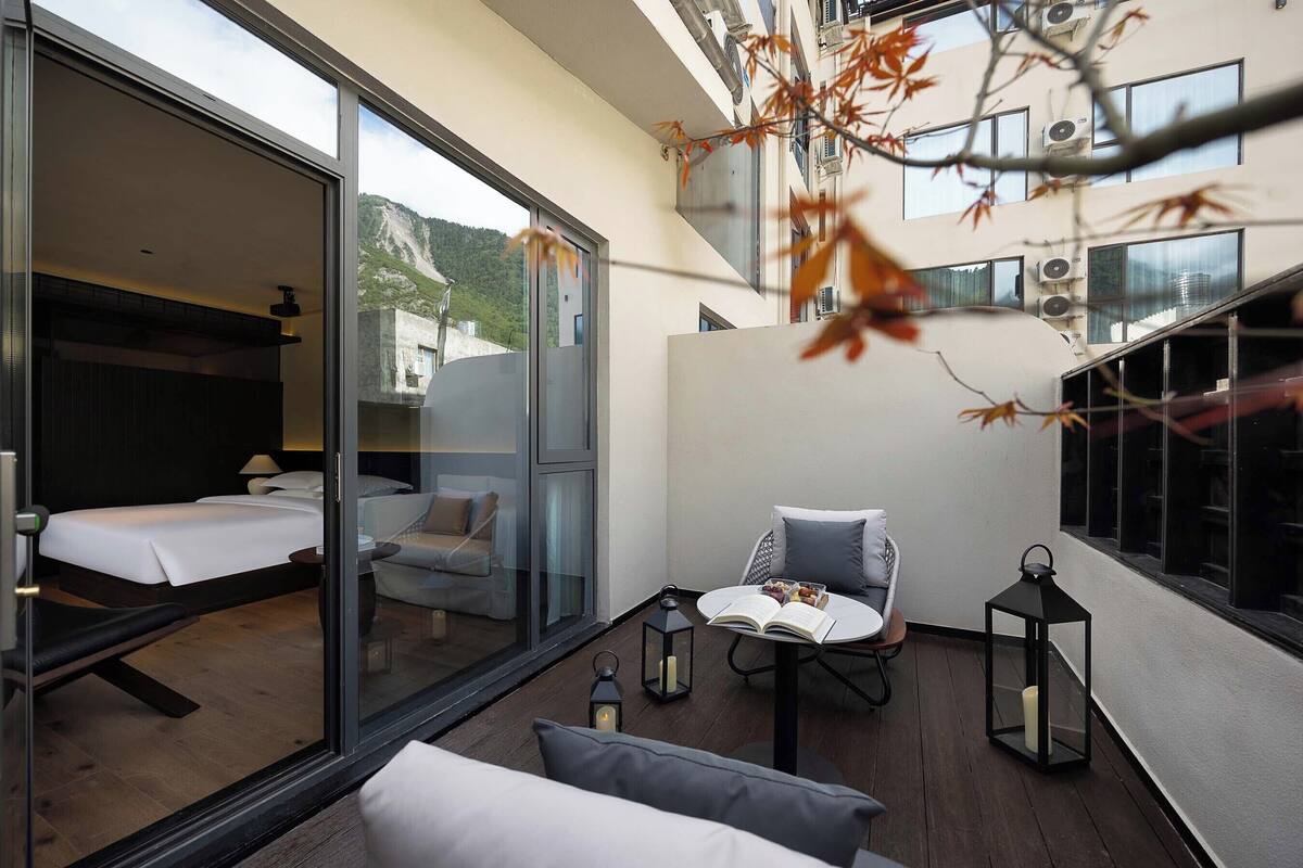 A modern hotel room with a glass wall, a balcony with seating, and a view of the mountains.