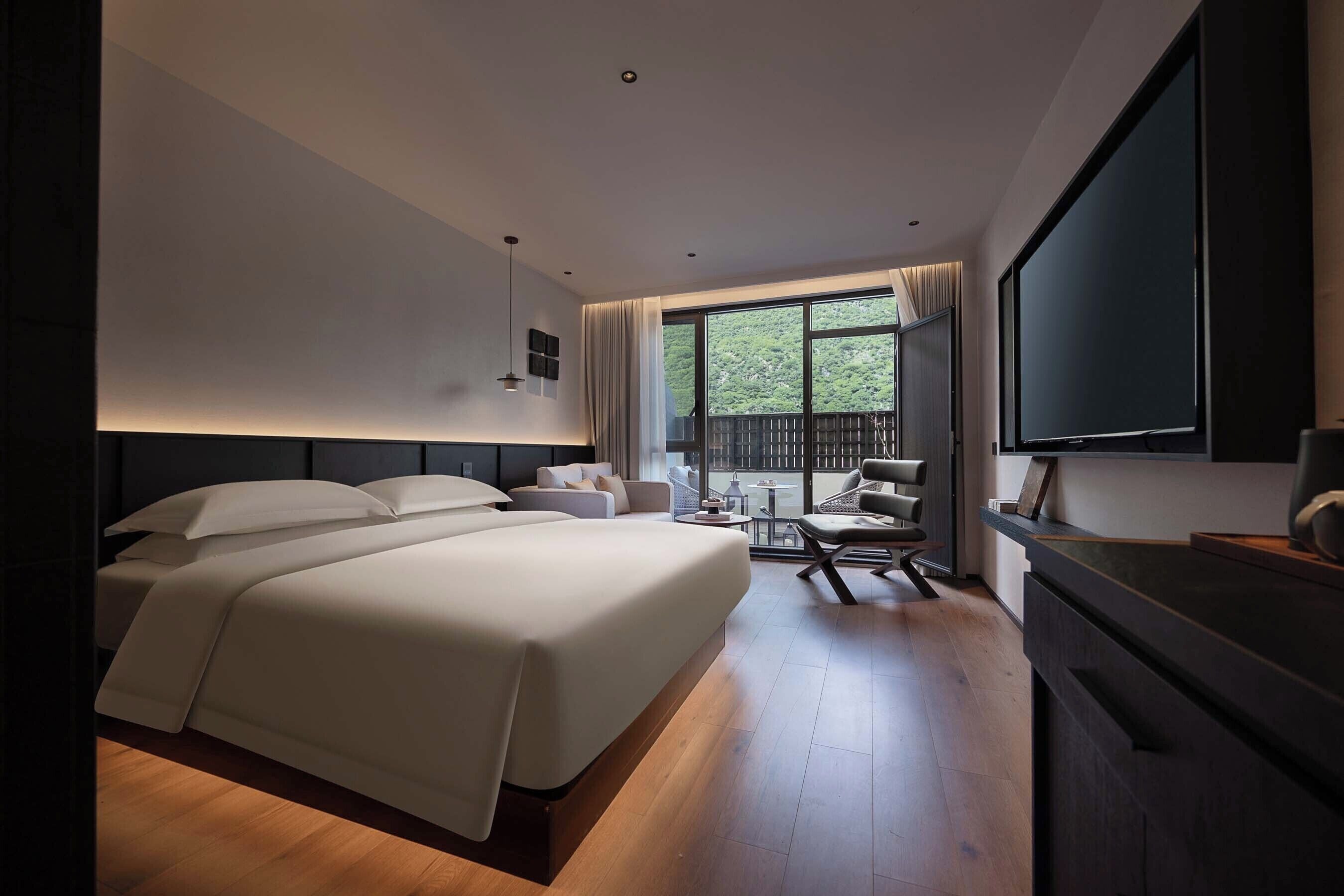 Una habitación de hotel moderna con una cama grande, televisión, balcón con vista a zonas verdes y un comedor con mesa y sillas.