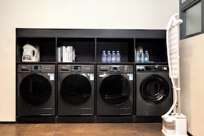 Une buanderie équipée de quatre machines à laver, d’un chauffe-eau et d’une étagère où sont rangés des livres et des bouteilles d’eau.