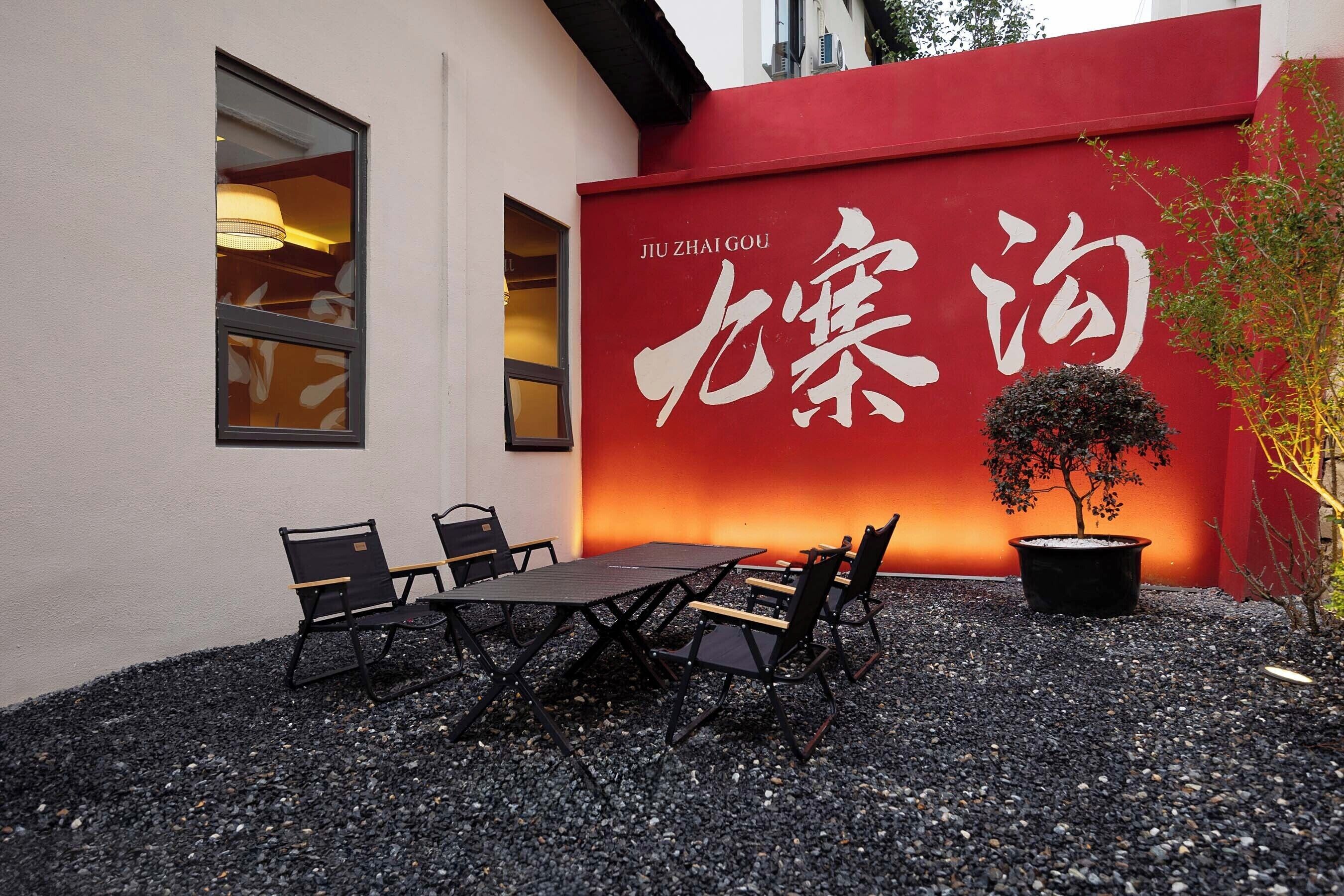 An outdoor seating area with a table and chairs, a potted plant, and a red wall with white Chinese characters.