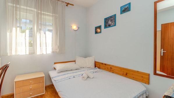 Ein Schlafzimmer mit einem Holz-Kopfteil, einem Einzelbett mit weißer Bettwäsche, einem Nachttisch, einer Holzkommode und einem Fenster mit transparenten Vorhängen.