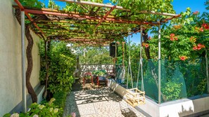 Ein überdachter Gartenbereich mit einer Pergola, Kletterpflanzen, einer Schaukel und einer Sitzgelegenheit.