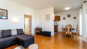 Ein Wohnzimmer mit einer Couch, Hocker und einem kleinen Tisch. Es gibt eine Küchenzeile mit einem Esstisch und Stühlen.