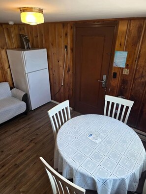 A room with a round table covered with a white tablecloth, two white chairs, a white refrigerator, and a wooden door. - Unwind Steps from the Ocean in Coastal Serenity in North Truro, Massachusetts (North Truro)