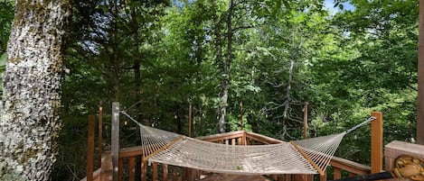 Una cadira l'hamaca penjada entre bigues de fusta sobre un pont de fusta envoltat d'arbres.