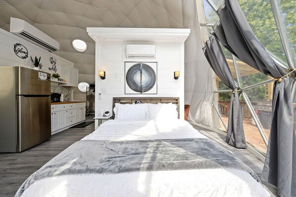 A modern bedroom with a large bed, a kitchen area, and a view through a window with curtains.