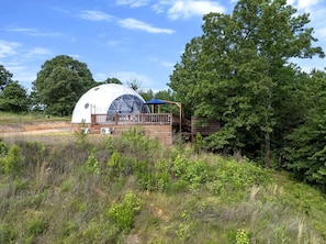 A dome-shaped structure with a wooden deck and a covered area, surrounded by greenery and trees.