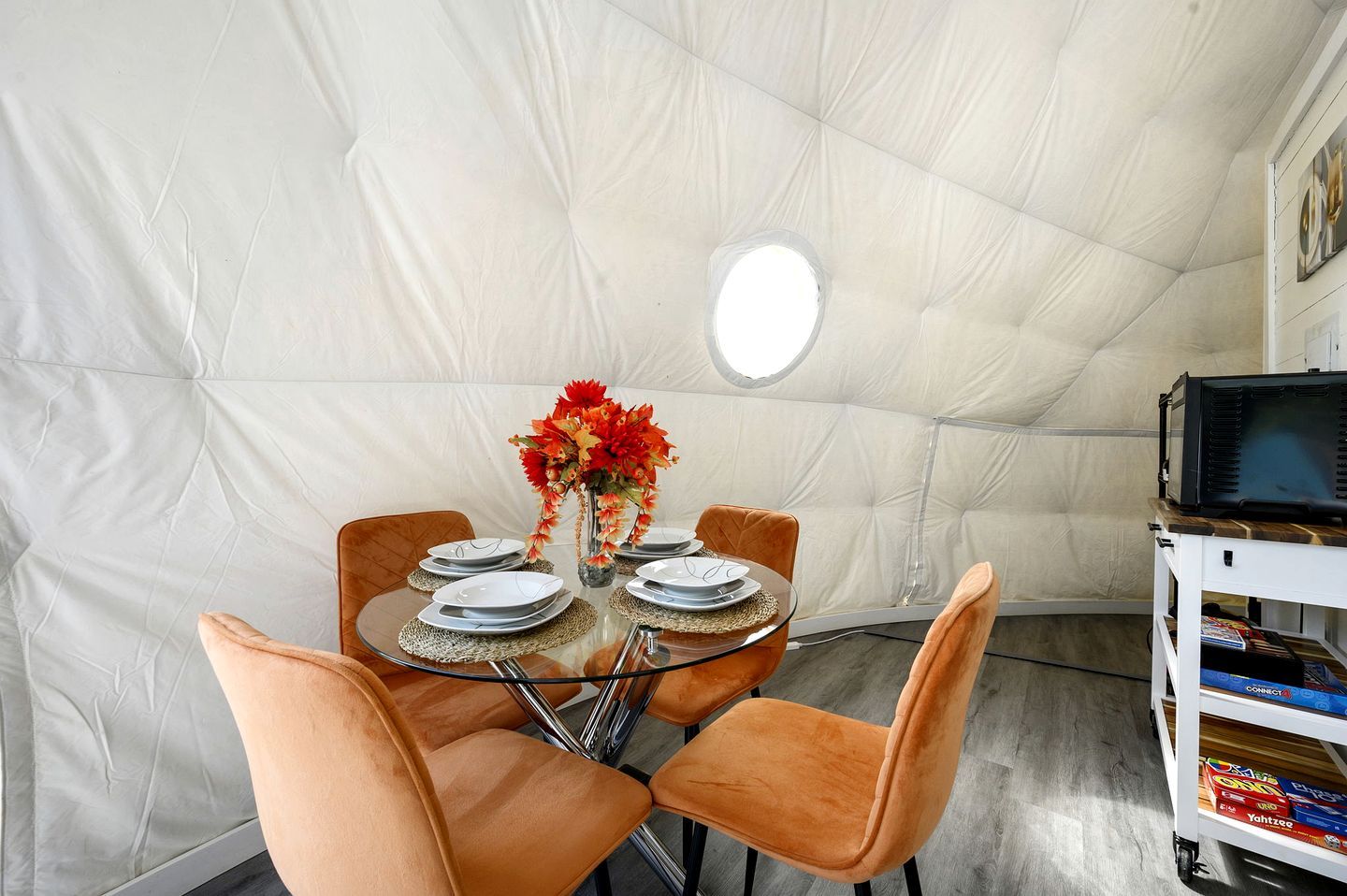 A dining area with a round glass table, orange chairs, and a white tent-like structure in the background.
