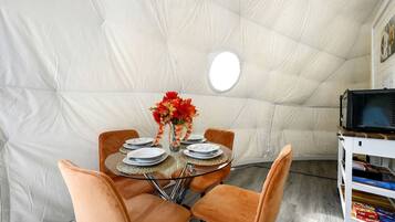 A dining area with a round glass table, orange chairs, and a white tent-like structure in the background.