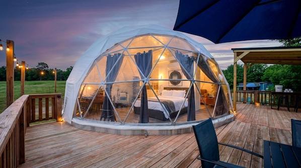 Une structure en forme de dôme géodésique abritant un lit et un espace salon, entourée d’une terrasse en bois et d’un éclairage extérieur.