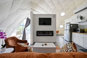 A tented accommodation with a flat-screen TV, fireplace, dining area, kitchen, and a view through the window.
