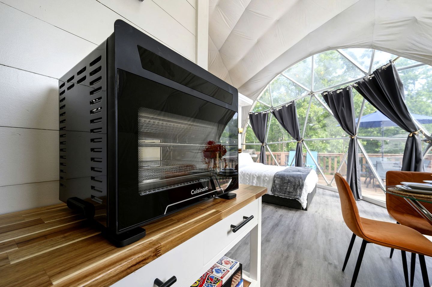 Une pièce moderne avec un grand four noir, un lit recouvert d’une couverture blanche et grise, une table à manger avec des chaises, et une grande fenêtre avec des rideaux.