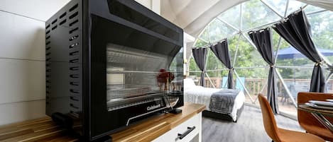 Une pièce moderne avec un grand four noir, un lit recouvert d’une couverture blanche et grise, une table à manger avec des chaises, et une grande fenêtre avec des rideaux.