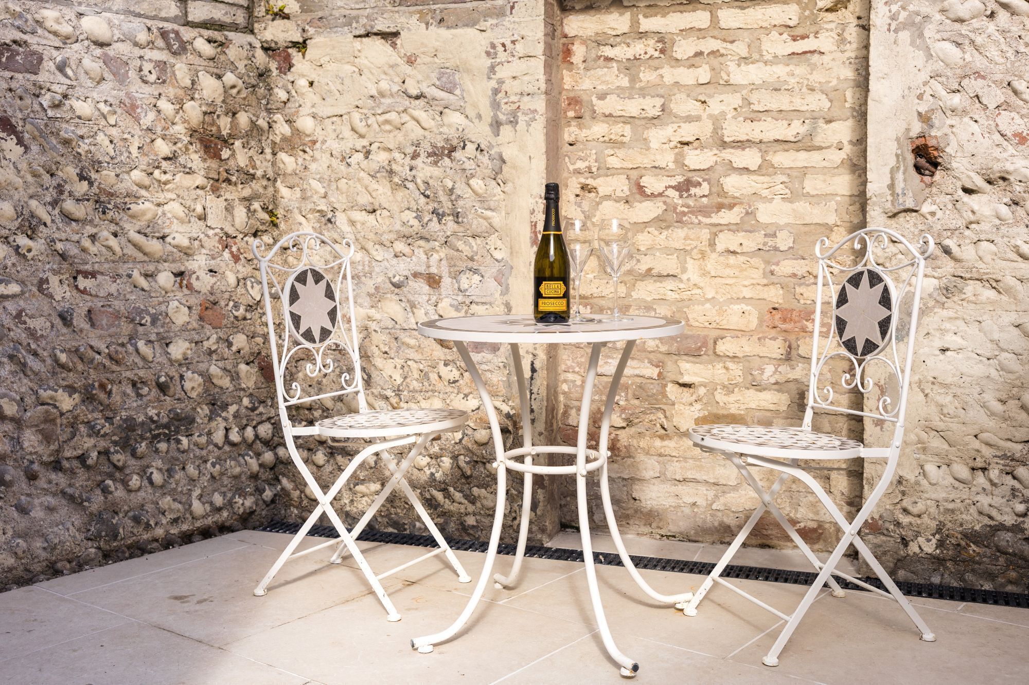 Ein Set aus weißem Metalltisch und -stühlen vor einer Steinwand. Auf dem Tisch steht eine Flasche Prosecco.