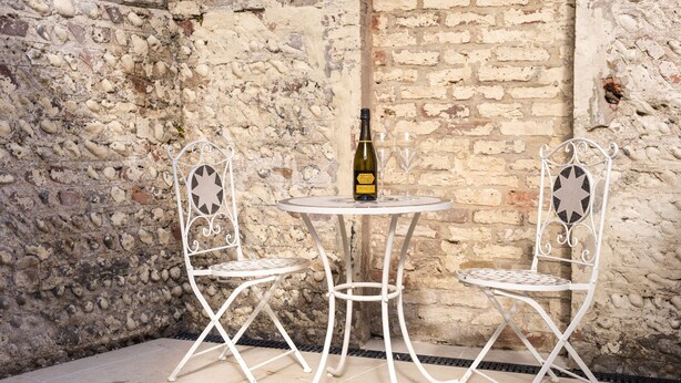 A white metal table and chairs set against a stone wall. A bottle of Prosecco wine is placed on the table. - Little Meads House - charming cottage close to seafront (Eastbourne)