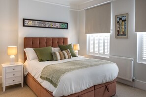 Ein Schlafzimmer mit einem Bett, Nachttischen, einem Heizkörper und einem Fenster mit Jalousien.