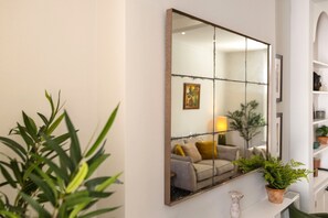 Ein Zimmer mit einem großen Spiegel, der ein Sofa und eine Lampe reflektiert. Auf einem Regal steht eine Pflanze und auf dem Boden eine weiße Vase.