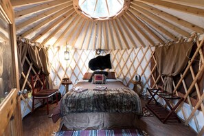 A yurt interior with a bed, a fur rug, a wooden chair, and a hanging lantern.