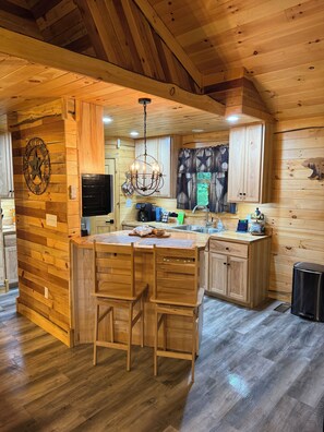 A wooden cabin interior with a kitchen island, bar stools, a chandelier, and a clock on the wall. - 2 Bedroom upper Barndominium cabin, Hot tub, King bed,Fire Pit, Horses on site

 (Logan)