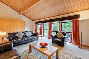 Ein geräumiges Wohnzimmer mit Holzbalkendecke, Ledersofas, einem Couchtisch und großen Fenstern mit Vorhängen.