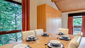 Een eetkamer met een houten tafel gedekt voor zes personen, grote ramen met gordijnen, en een houten plafond.