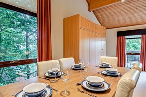 Ein Esszimmer mit Holztisch, gedeckten Plätzen für sechs Personen, großen Fenstern mit Vorhängen und einer Holzbalkendecke.