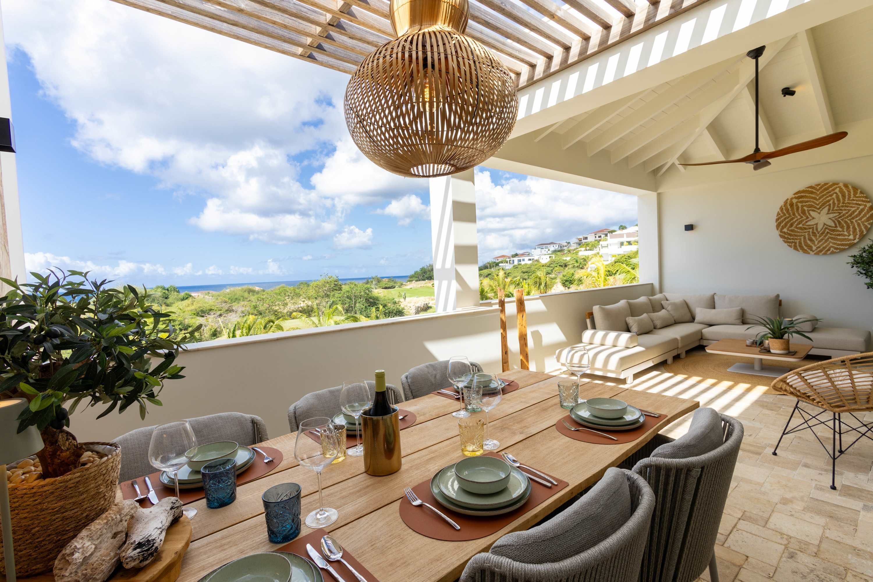 Un coin repas avec une table dressée pour six personnes, un ventilateur de plafond en osier et une vue sur la mer.