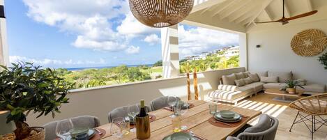 Un coin repas avec une table dressée pour six personnes, un ventilateur de plafond en osier et une vue sur la mer.