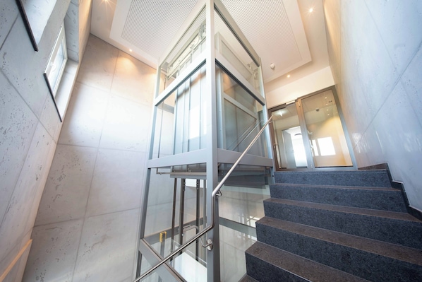 A modern interior with a glass elevator, marble flooring, and a staircase. - Hotel BoxenStop (Dormagen)