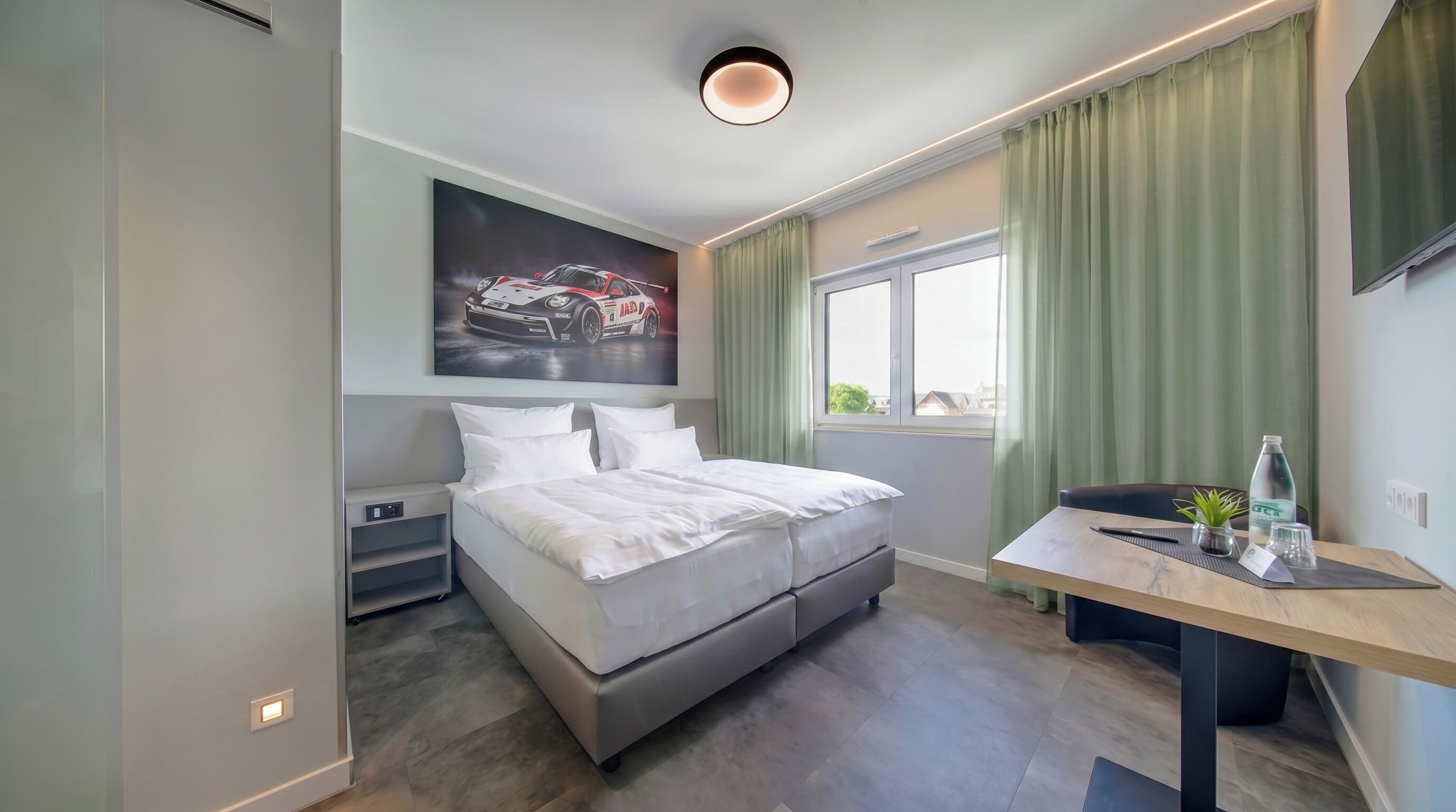 A modern hotel room with a bed, a desk, a window with curtains, a wall-mounted artwork, and a ceiling light.