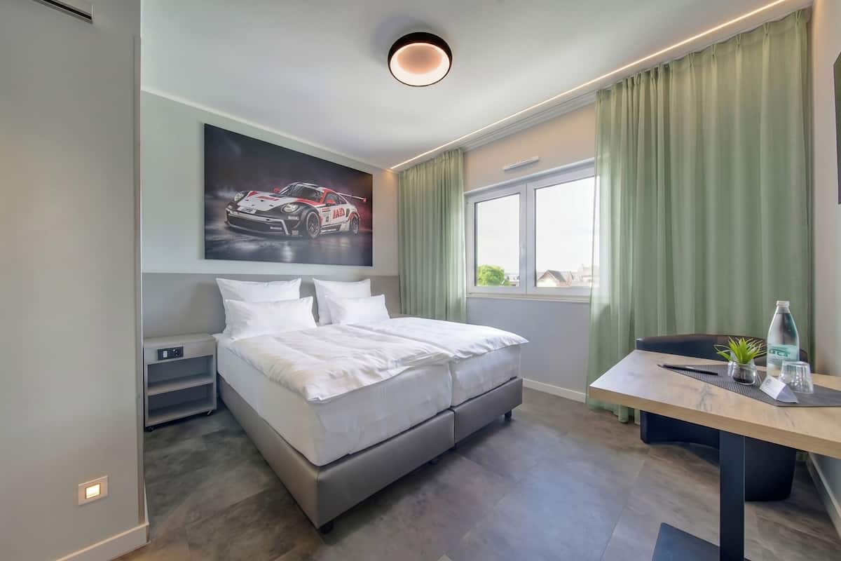 A modern hotel room with a bed, a desk, a window with curtains, a wall-mounted artwork, and a ceiling light.