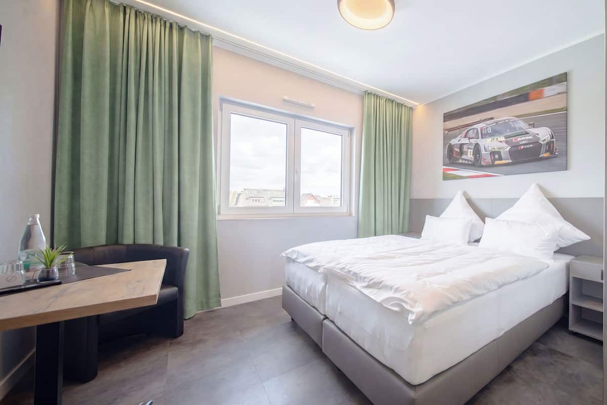 A hotel room with a bed, a desk, a chair, a window, and a painting on the wall.