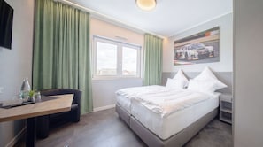 Ein Hotelzimmer mit Bett, Schreibtisch, Stuhl, Fenster und einem Bild an der Wand.