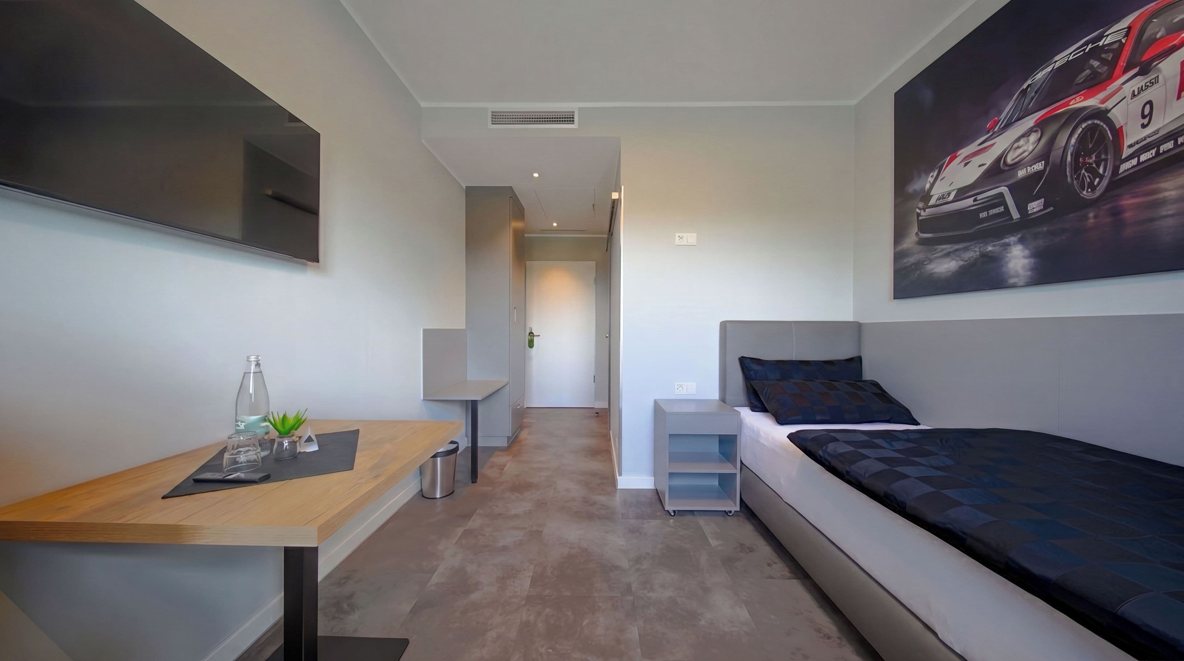 A modern bedroom with a bed, a desk, a chair, a small table with a plant, and a wall-mounted TV.