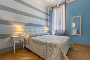 Ein Schlafzimmer mit einer gestreiften Wand, einem Bett mit floralem Überwurf, einem Nachttisch mit Lampen und einem Spiegel an der Wand.