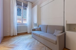 Ein modernes Wohnzimmer mit einer Couch, einem Heizkörper, einem Fenster mit Vorhängen und einem Fischgrätenparkettboden.