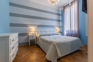 Een slaapkamer met een gestreepte wand, een bed met een beddegoed met dessin, een nachtkastje met een lamp en een kapstok.