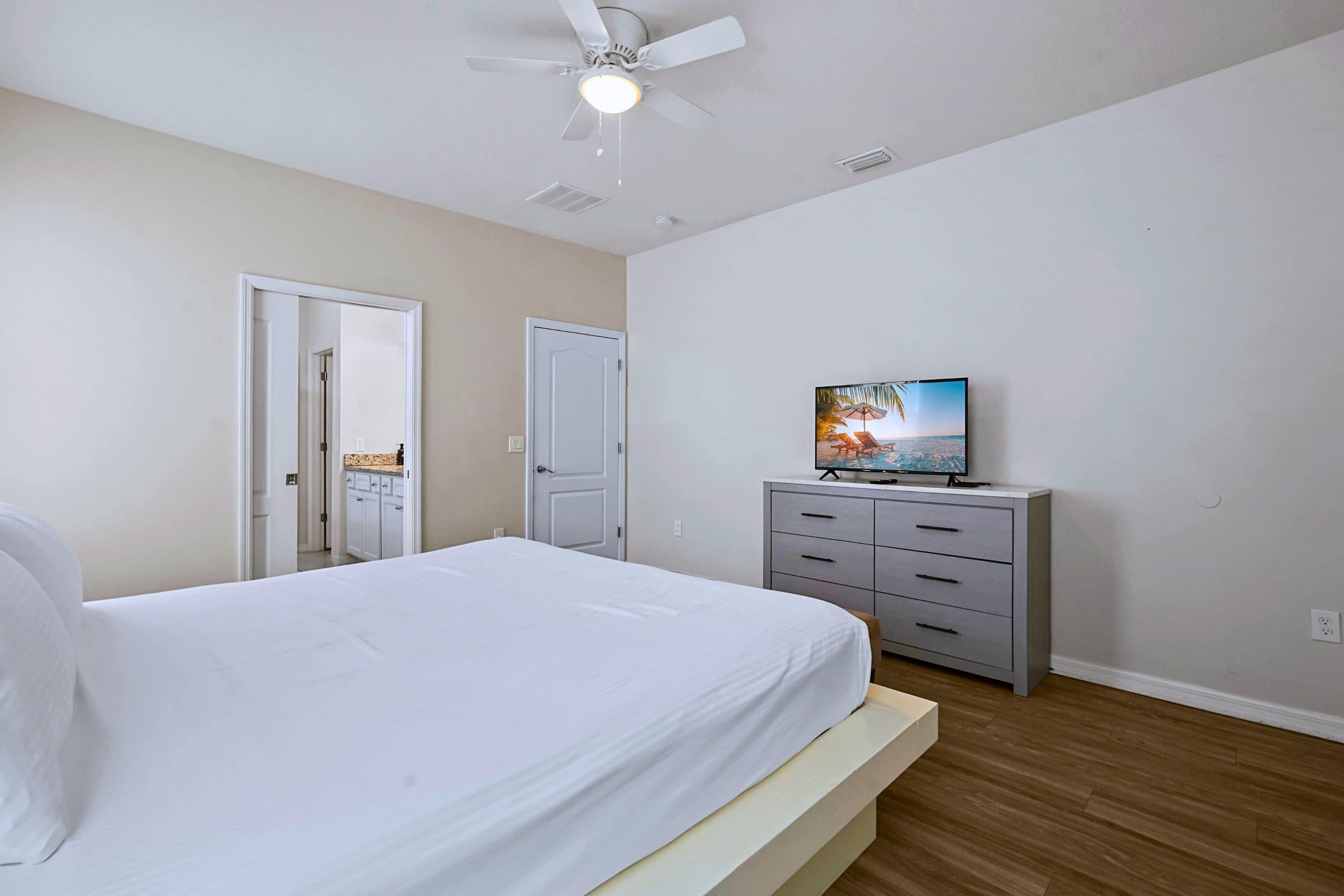 Une chambre à coucher avec un lit, un ventilateur de plafond, un téléviseur posé sur une commode et une porte donnant sur une autre pièce.