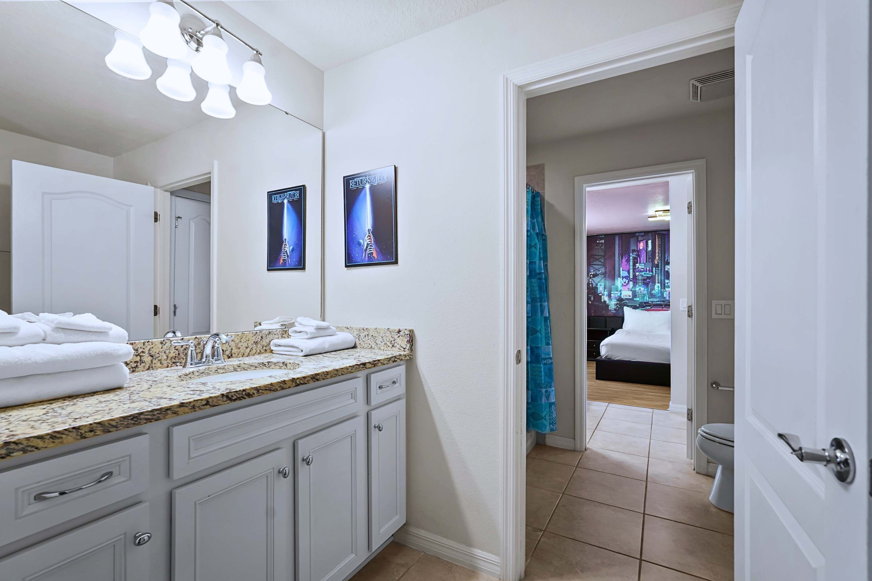 Une salle de bain avec un comptoir en granit, des armoires blanches et un grand miroir. Deux affiches encadrées sont accrochées au mur, et un rideau de douche bleu est visible à l’arrière-plan.