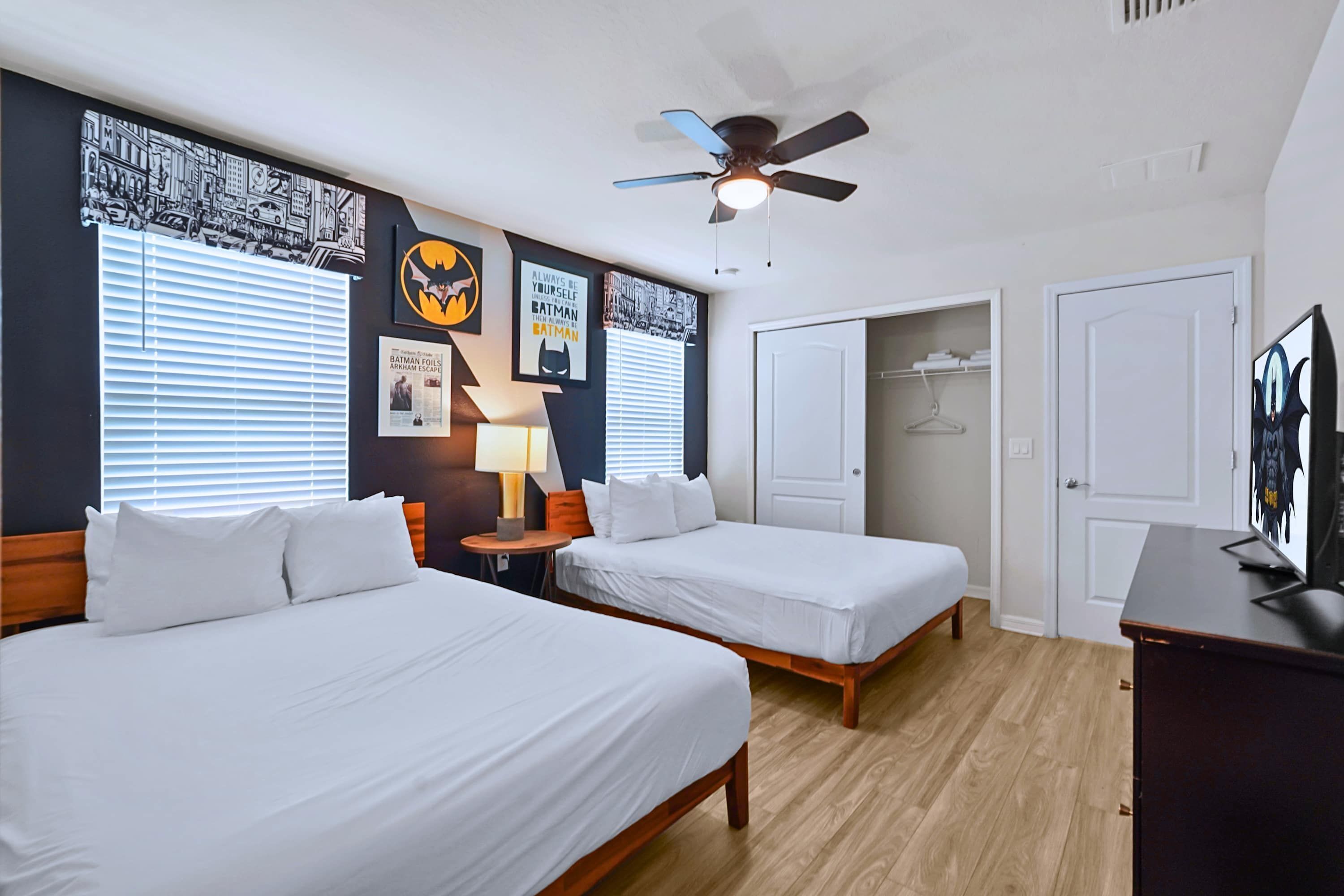 A hotel room with two beds, a ceiling fan, a wall with Batman-themed artwork, and a door leading to a closet.